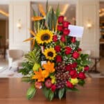 Arreglo floral premium con girasoles, rosas rojas, lirios, uvas, manzanas, peras y una botella de vino, sobre una mesa de mad