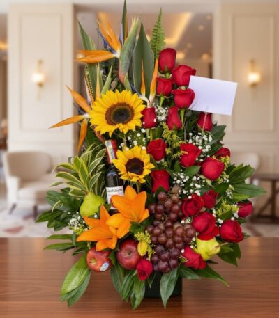 Arreglo floral premium con girasoles, rosas rojas, lirios, uvas, manzanas, peras y una botella de vino, sobre una mesa de mad