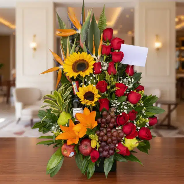 Arreglo floral premium con girasoles, rosas rojas, lirios, uvas, manzanas, peras y una botella de vino, sobre una mesa de mad