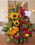Arreglo floral premium con girasoles, rosas rojas, lirios, uvas, manzanas, peras y una botella de vino, sobre una mesa de mad