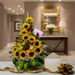 Arreglo floral de girasoles y rosas rojas con frutas frescas sobre mármol, con cinta dorada y lazo, en un elegante recibidor.