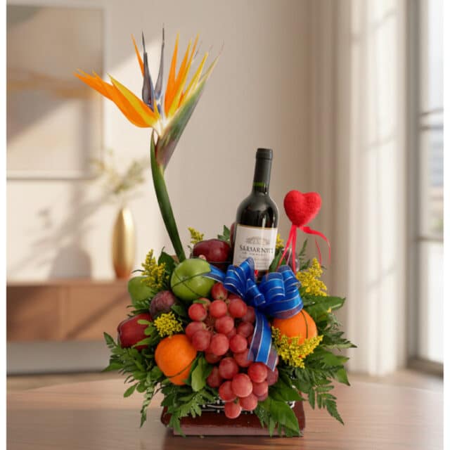 Arreglo floral gourmet ITALINA con ave del paraíso, vino tinto, uvas, manzanas, naranjas y ciruelas, sobre una mesa de madera