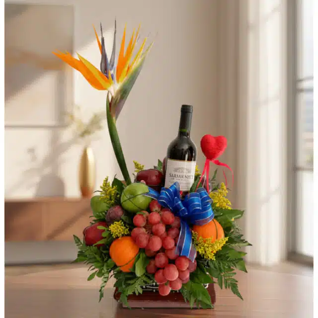 Arreglo floral gourmet ITALINA con ave del paraíso, vino tinto, uvas, manzanas, naranjas y ciruelas, sobre una mesa de madera