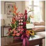 Arreglo floral PIA con rosas, lirios, heliconias y aves del paraíso, sobre una mesa de madera, con manos colocando una tarjet