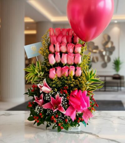 Arreglo floral premium 'Quiero que Sepas' con rosas rosadas, lirios, follaje verde y un globo fucsia, sobre mármol en un lobb