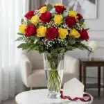 Arreglo floral LORENZA con 24 rosas rojas y amarillas, follaje verde, y gypsophila en un jarrón de cristal, acompañado de una