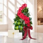Arreglo floral alto con rosas rojas voluta, follaje verde, y pequeñas flores blancas, en un jarrón de cristal con un lazo de