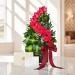 Arreglo floral alto con rosas rojas voluta, follaje verde, y pequeñas flores blancas, en un jarrón de cristal con un lazo de