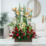 Arreglo floral 'Siempre Juntos' con aves del paraíso, lirios blancos y rosas rojas en base de madera, sobre mármol.