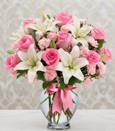 Arreglo floral vibrante con rosas rosadas, lirios blancos y claveles rosados en un jarrón de cristal con lazo rosa, ideal par