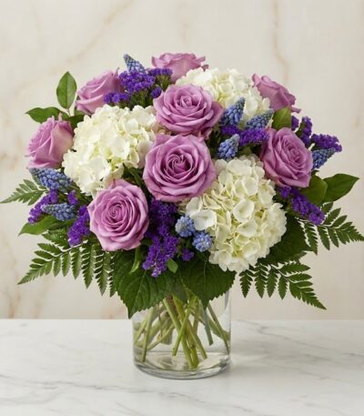 Arreglo floral VITTORIA con rosas lilas, hortensias blancas y muscari azul en un jarrón de cristal, sobre una mesa de mármol.