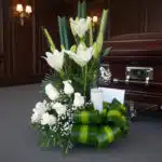 Arreglo floral fúnebre Claridad con lilas y rosas blancas, follaje verde, vela y tarjeta, junto a un ataúd en sala de velació