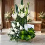 Arreglo fúnebre Claridad con lirios y rosas blancas, vela, follaje verde, en primer plano con fondo de sala de velación premi