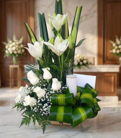 Arreglo fúnebre Claridad con lirios y rosas blancas, vela, follaje verde, en primer plano con fondo de sala de velación premi