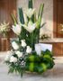 Arreglo fúnebre Claridad con lirios y rosas blancas, vela, follaje verde, en primer plano con fondo de sala de velación premi