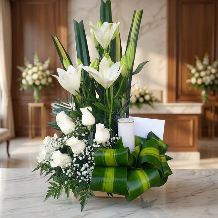 Arreglo fúnebre Claridad con lirios y rosas blancas, vela, follaje verde, en primer plano con fondo de sala de velación premi
