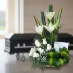 Arreglo floral fúnebre Claridad con lirios y rosas blancas, vela y tarjeta, en sala de velación moderna con ataúd al fondo.