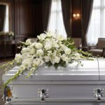 Arreglo fúnebre cubre caja Asunción con lirios blancos, claveles y follaje verde sobre un ataúd gris en una sala de velación
