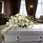 Arreglo fúnebre cubre caja Asunción con lirios blancos, claveles y follaje verde sobre un ataúd gris en una sala de velación