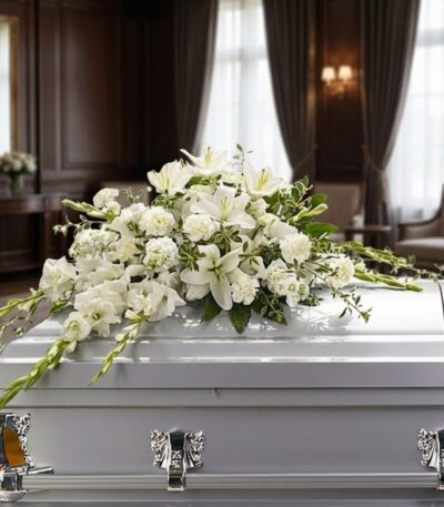 Arreglo fúnebre cubre caja Asunción con lirios blancos, claveles y follaje verde sobre un ataúd gris en una sala de velación