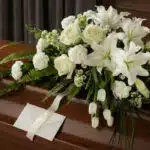 Arreglo fúnebre cubre caja Bondad con lirios, rosas y claveles blancos, follaje verde, cinta satinada y sobre de condolencias