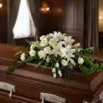 Arreglo floral fúnebre cubre caja 'Bondad' con lirios, rosas y claveles blancos, sobre un ataúd de madera oscura en una sala