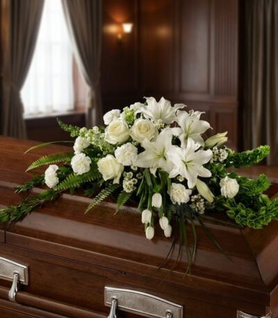 Arreglo floral fúnebre cubre caja 'Bondad' con lirios, rosas y claveles blancos, sobre un ataúd de madera oscura en una sala
