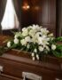 Arreglo floral fúnebre cubre caja 'Bondad' con lirios, rosas y claveles blancos, sobre un ataúd de madera oscura en una sala