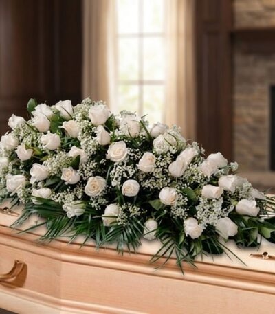 Arreglo fúnebre cubre caja con rosas blancas y follaje verde, sobre un ataúd de madera clara en una sala de velación premium