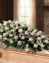 Arreglo fúnebre cubre caja con rosas blancas y follaje verde, sobre un ataúd de madera clara en una sala de velación premium