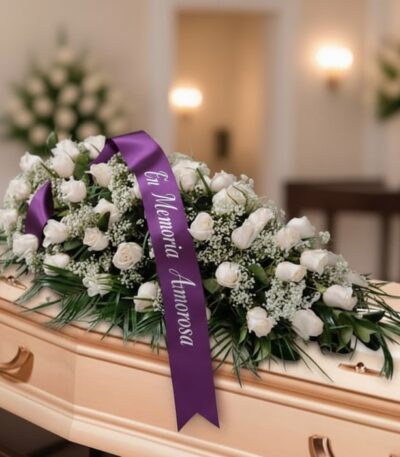 Arreglo fúnebre cubre caja Cielo con rosas blancas y follaje verde, adornado con una cinta morada que dice 'En Memoria Amoros