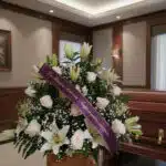 Arreglo floral fúnebre 'Infinito' con rosas blancas, lirios y gypsophila, cinta morada con mensaje de condolencia, en una sal
