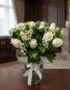 Arreglo fúnebre Pétalos del Alma con rosas blancas y follaje verde en un jarrón de cristal con lazo blanco, sobre una mesa de