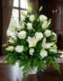 Arreglo fúnebre Piedad con rosas y lirios blancos, hojas verdes, en sala de velación premium con luz natural y bokeh.