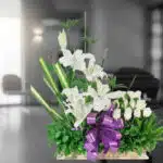 Arreglo fúnebre Pureza con lirios y rosas blancas, lazo morado, en sala de velación moderna con ataúd desenfocado al fondo.