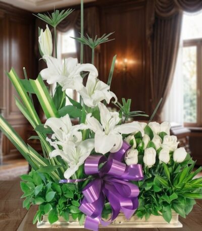 Arreglo fúnebre Pureza con lirios y rosas blancas, lazo morado, en sala de velación premium con luz natural y bokeh.