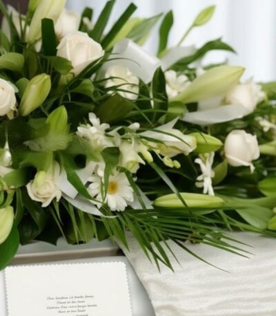 Arreglo fúnebre cubre caja con rosas blancas, lirios y follaje verde, con una cinta satinada blanca y una tarjeta de condolen