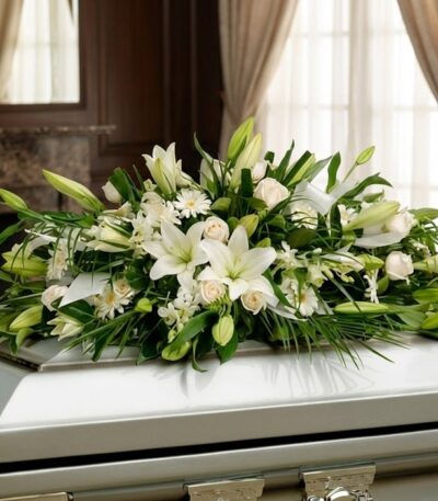 Arreglo fúnebre cubre caja con rosas blancas, lirios y follaje verde, presentado en una sala de velación premium con luz natu
