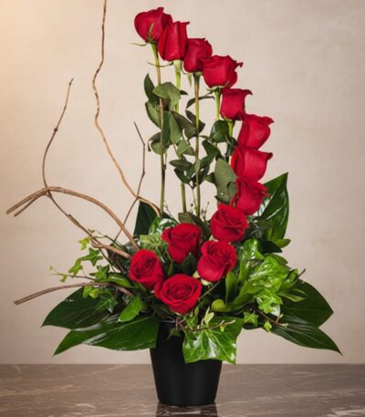 Arreglo floral GIANNA con 13 rosas rojas frescas en un jarrón negro, ramas decorativas y follaje verde, sobre una superficie