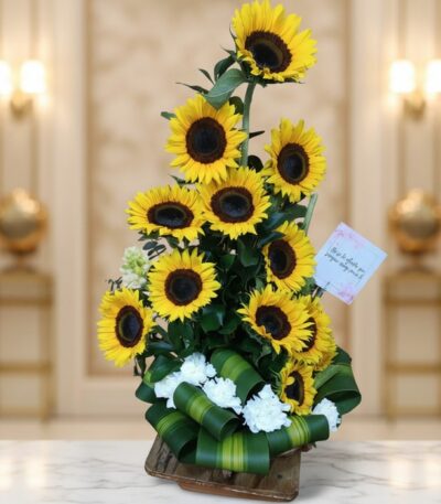 Arreglo floral de girasoles Sunna en un jarrón de madera, con detalles de flores blancas y lazo verde, sobre una superficie d