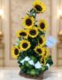 Arreglo floral de girasoles Sunna en un jarrón de madera, con detalles de flores blancas y lazo verde, sobre una superficie d