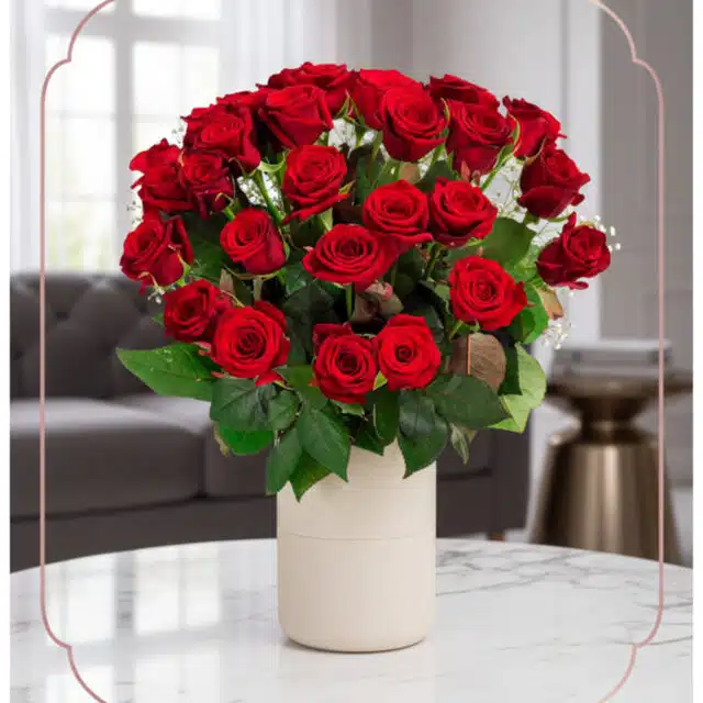 Arreglo floral Jianna con 25 rosas rojas frescas en un elegante jarrón blanco sobre mesa de mármol, fondo difuminado de sala