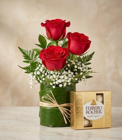 Elegante arreglo floral con tres rosas rojas vibrantes, follaje verde y flores blancas, acompañado de una caja de bombones Fe