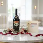 Botella de Baileys de 700ml rodeada de rosas y paniculata, con una tarjeta de regalo elegante y cinta de satén sobre una mesa