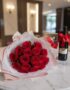 Elegante bouquet de 18 rosas rojas abiertas con empaque coreano, caja de chocolates y botella de vino tinto, sobre mesa de má