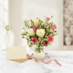 Bouquet Floral SUSANA con rosas blancas y rosadas, astromelias y follaje verde en jarrón de cristal, sobre mármol blanco con