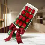 Caja de 12 rosas rojas frescas y vibrantes, presentadas en un elegante estuche blanco con un lazo de satén rojo, sobre una su
