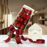 Caja de 12 rosas rojas frescas con lazo de satén rojo, tarjeta de regalo y cinta, sobre una mesa de mármol en un entorno eleg