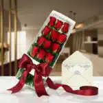 Caja de 12 rosas rojas frescas con lazo de satén rojo, tarjeta de regalo y cinta, sobre una mesa de mármol en un entorno eleg