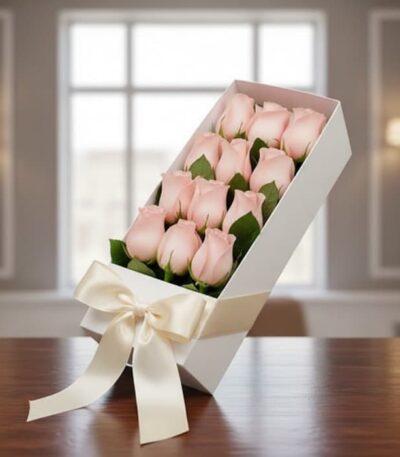 Caja blanca inclinada con 12 rosas rosadas frescas y un moño de satén, sobre una mesa de madera oscura con un fondo elegante